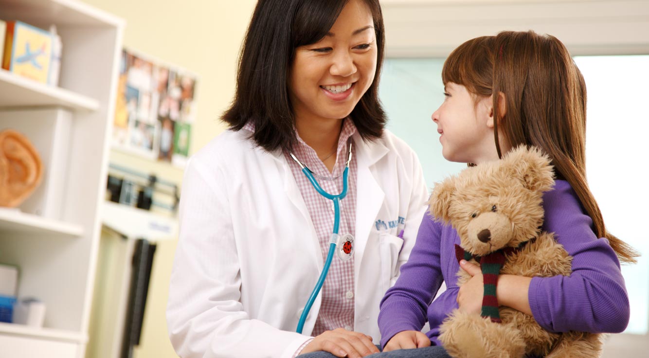 A little girl clutching a teddy bear smiles at her doctor.
