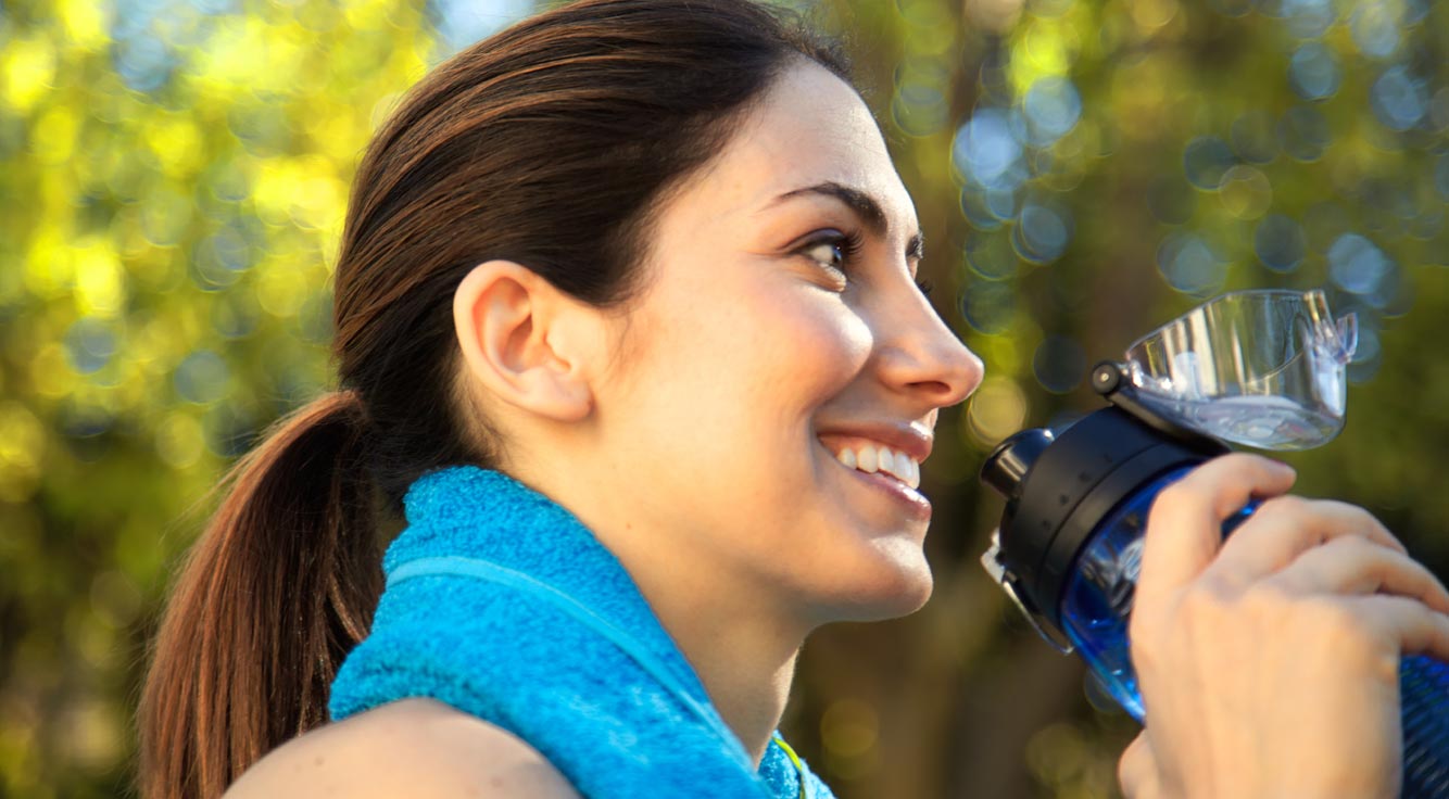 A smiling woman drinks water outdoors.