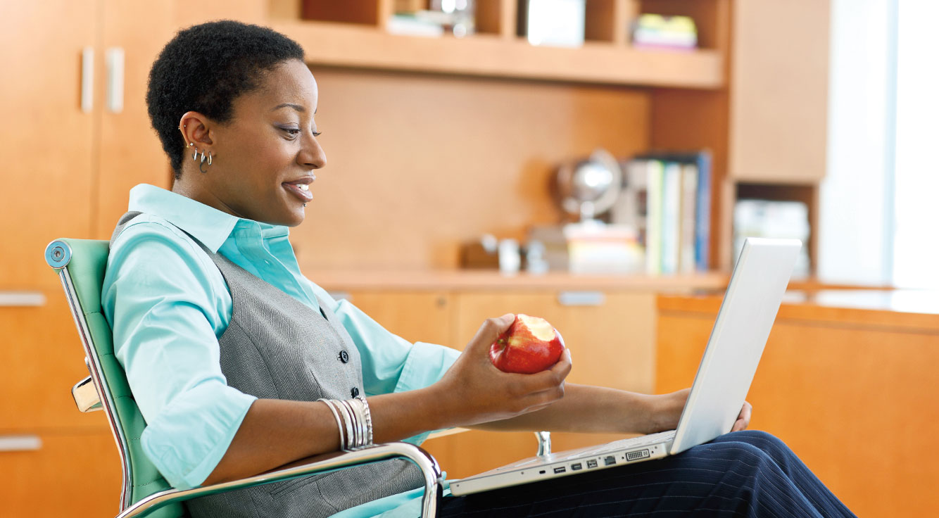A woman snacks on an apple as she faces a laptop.