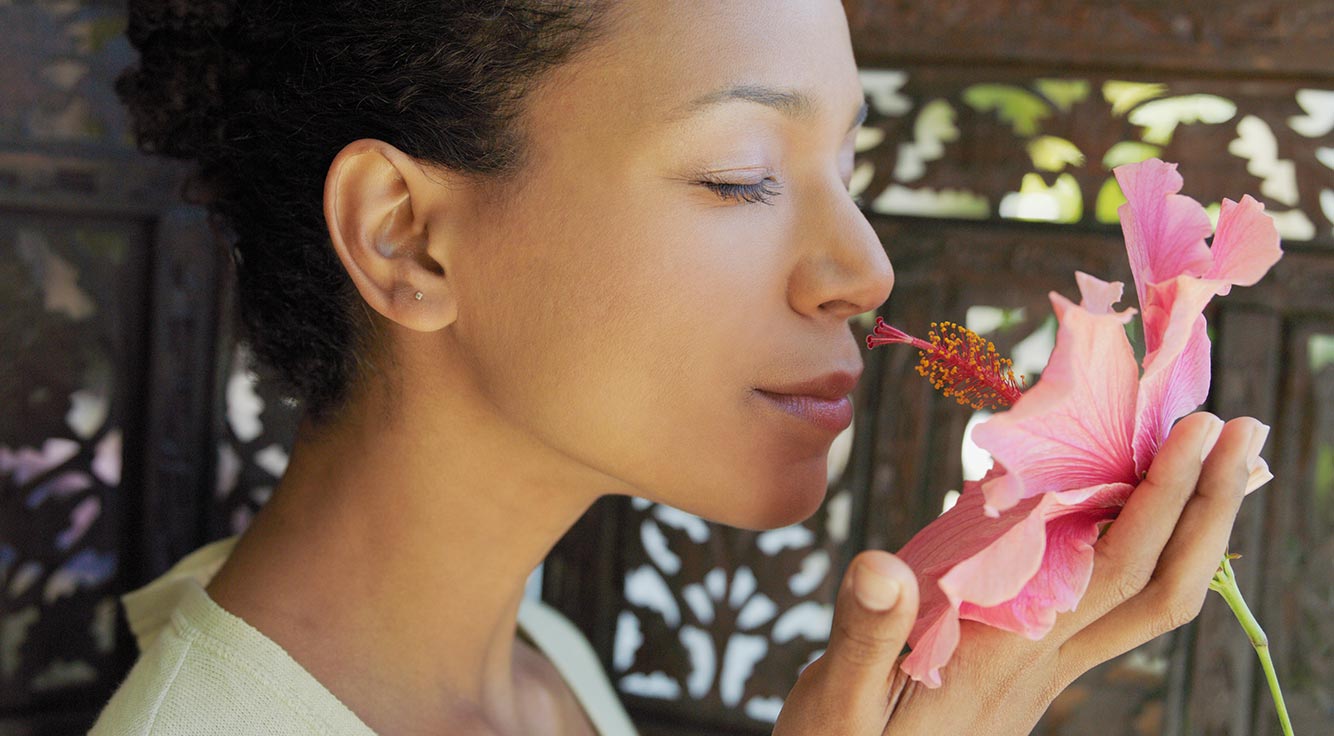 A woman in an outdoor spa setting with her eyes closed, smelling a hibiscus flower.
