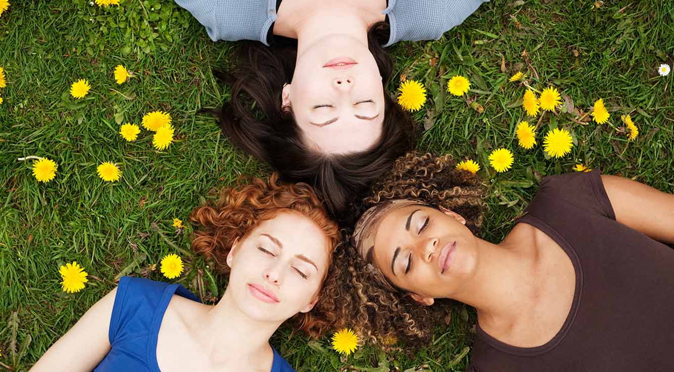Three young women relax on the grass with eyes closed while surrounded by yellow flowers