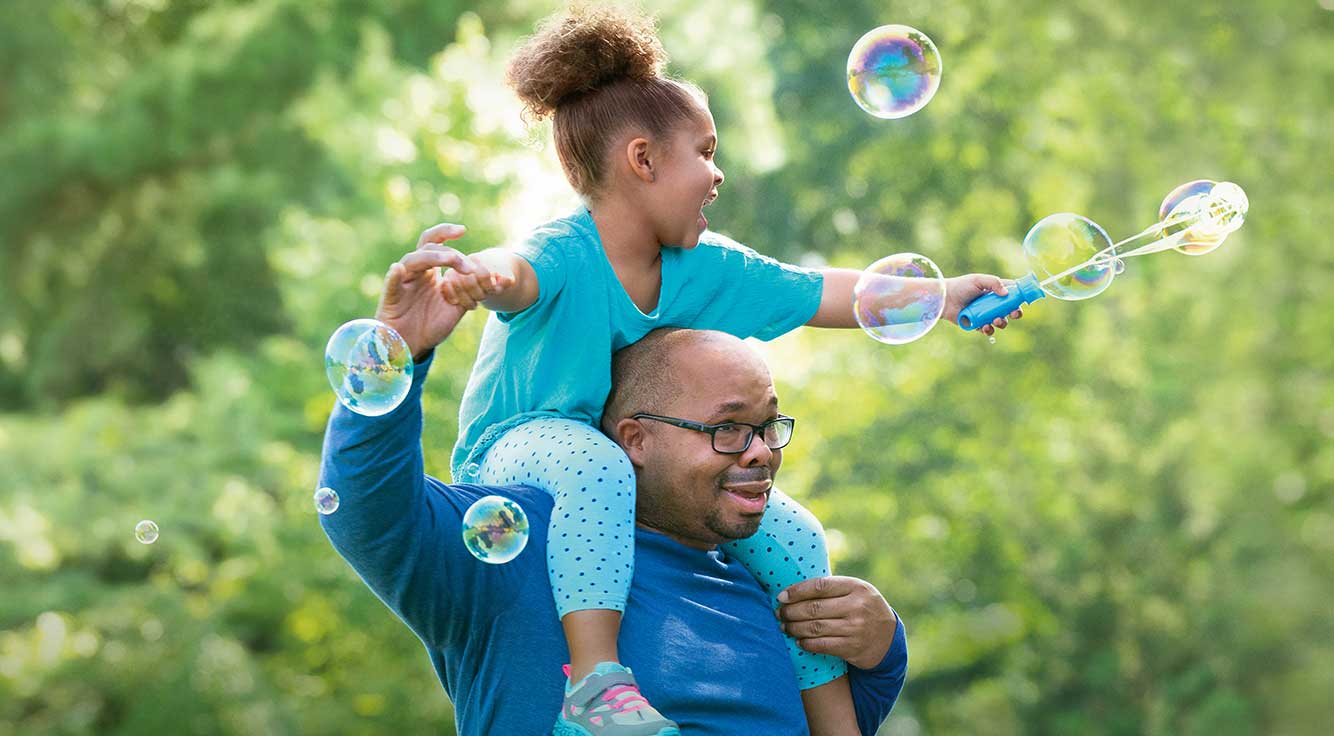 A young daughter playing with a bubble wand sits on her father’s shoulders.