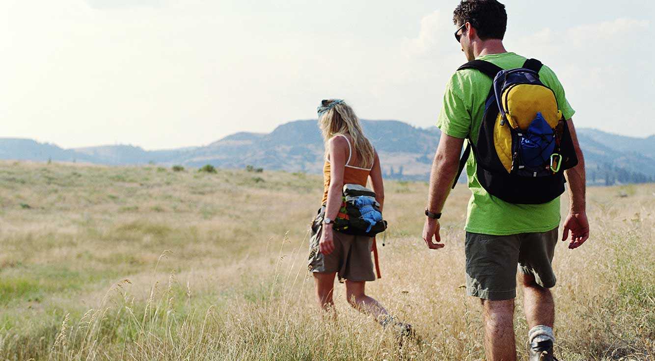 A couple hikes through a grassy field surrounded by mountains.
