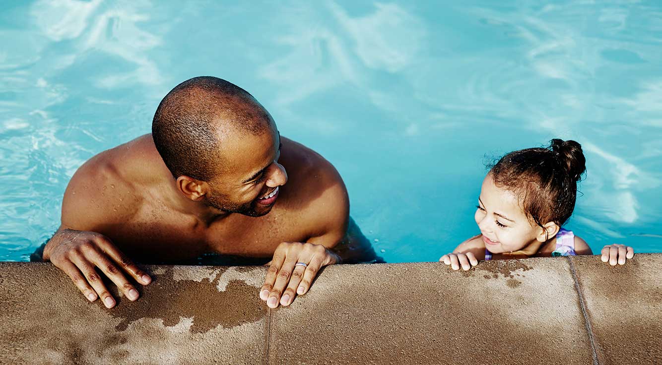 A father and his young daughter hold on to the side of the pool while smiling at each other.