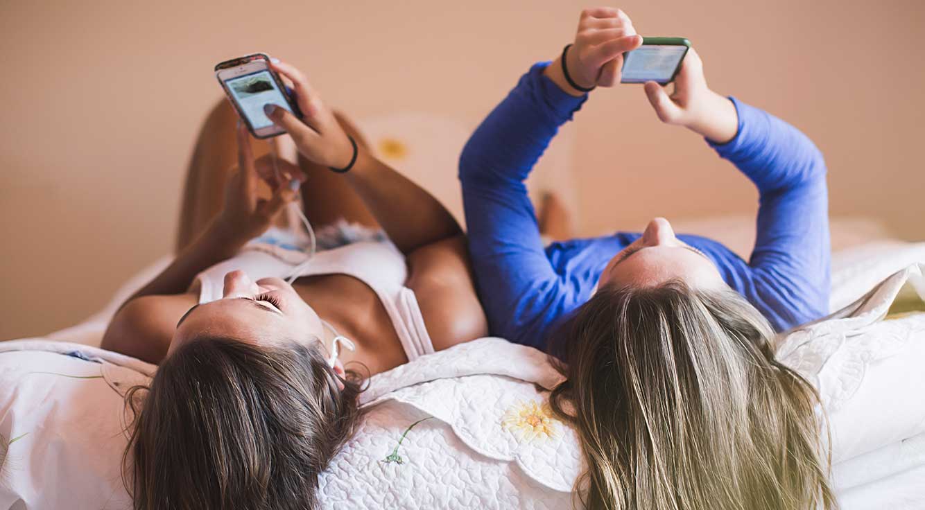 Two teen girls lay side by side on a bed while holding their smartphones up in the air