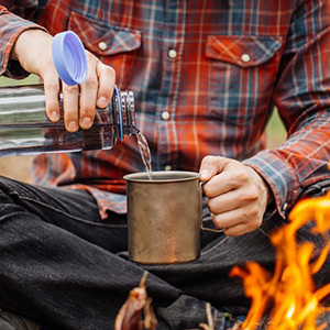 Man pours water into mug outside by a campfire..