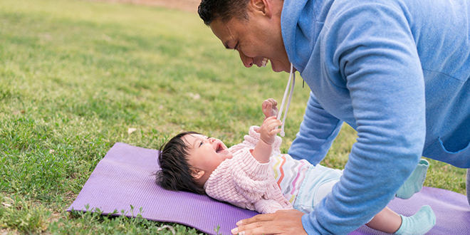 Father leaning over baby who is lying on yoga mat outside and laughing.