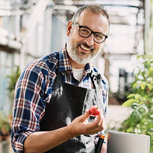An older man working in his indoor garden, holding a tomato and smiling.