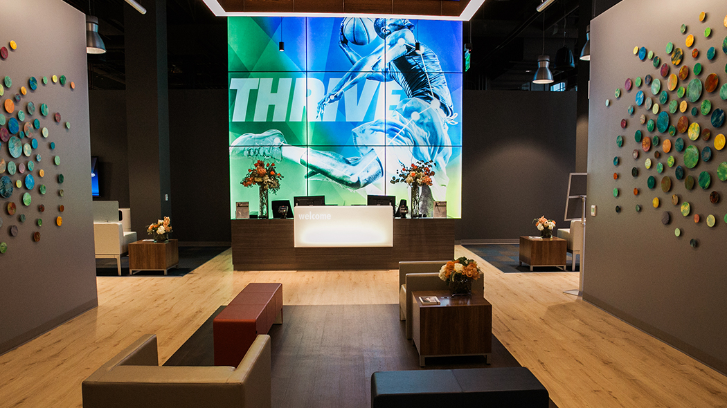Large modern reception area with blond wood floors and large lit up artwork of an athlete jumping with the word Thrive behind the desk
