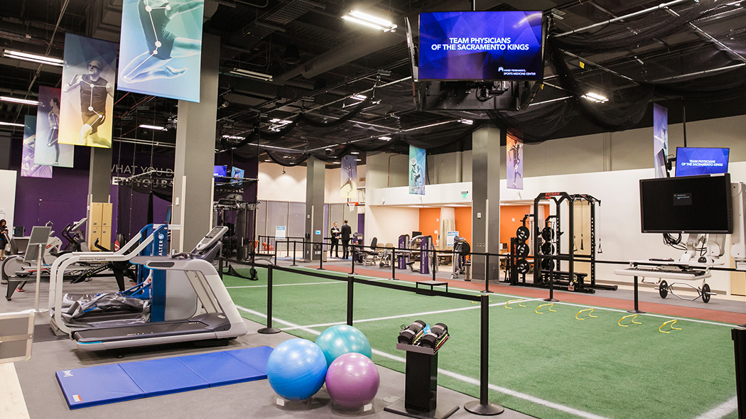 Rehabilitation open space and equipment room has images of athletes and TVs hanging from the ceiling; exercise machines, weights, mats and balls along the walls; and artificial turf running through the center